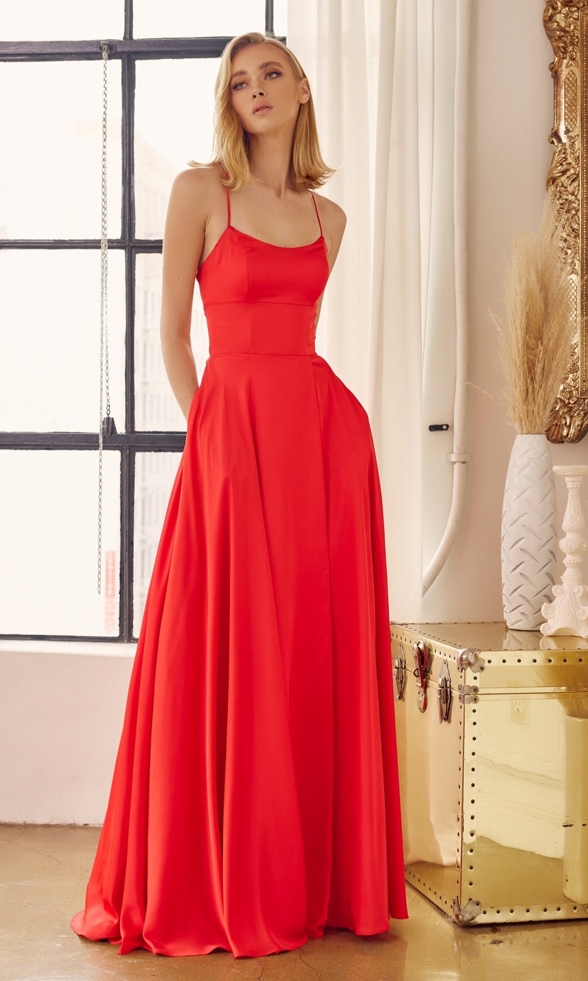 A woman stands indoors wearing the Strappy-Back Simple Long A-Line Prom Dress 278, a floor-length red gown with thin straps, posing near a window and gold trunk amid neutral decor.